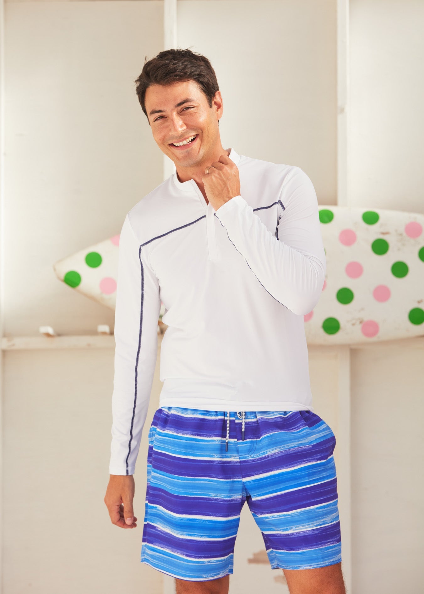 Man in front of a polka dotted surf board wearing the Cabana Life Sun Protective San Sebastian Stripe Men's Swim Trunk.