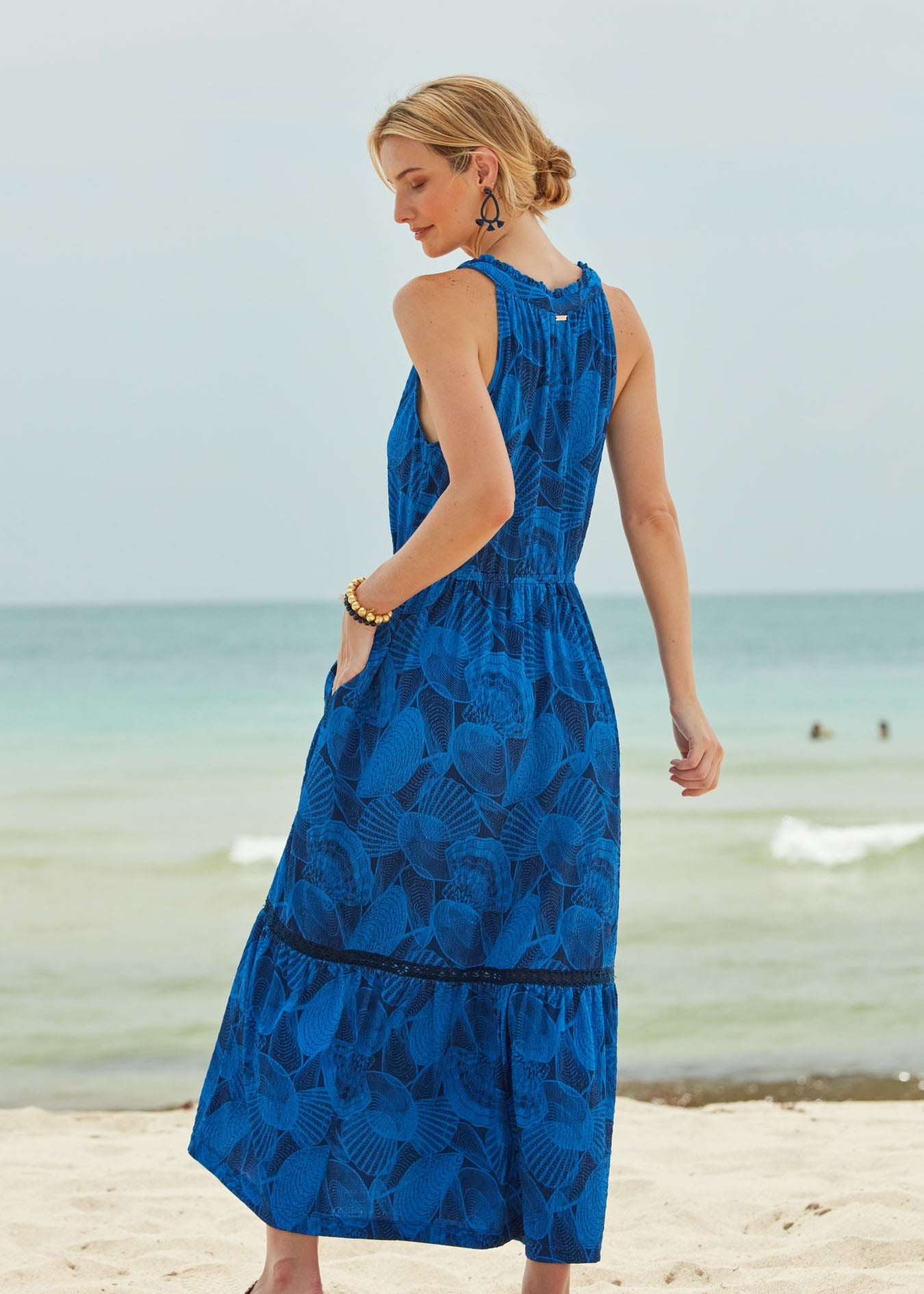 A blonde woman facing behind wearing the San Sebastian Tie Neck Maxi Dress on a beach.