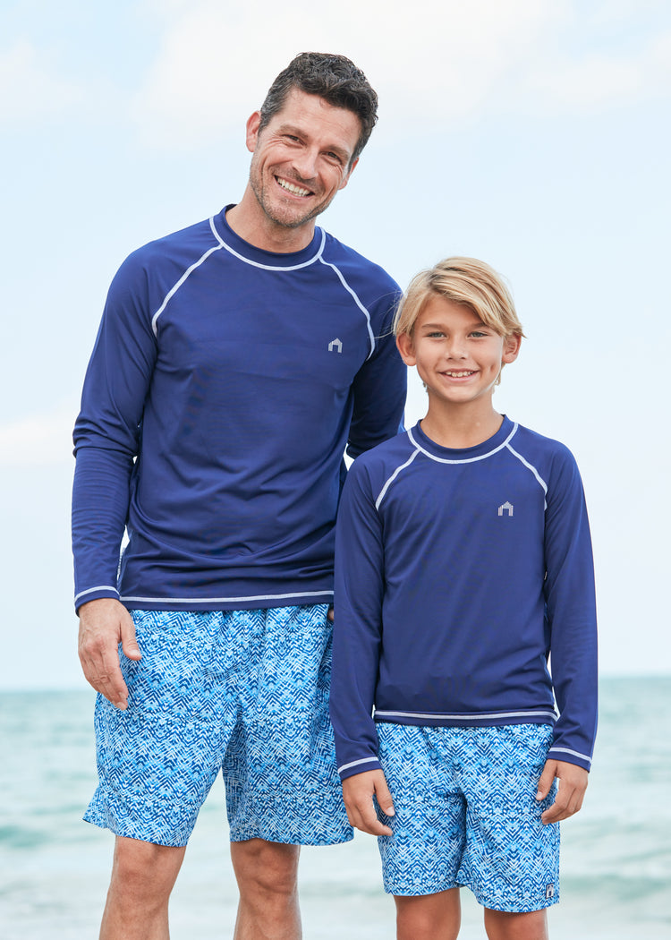 Man and boy by the ocean wearing matching Navy Long Sleeve Cabana Rashguard and Naples swim trunks. 