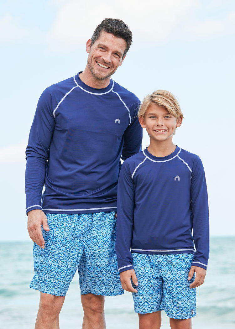 Man and boy by the ocean wearing matching Navy Long Sleeve Cabana Rashguard and Naples swim trunks.