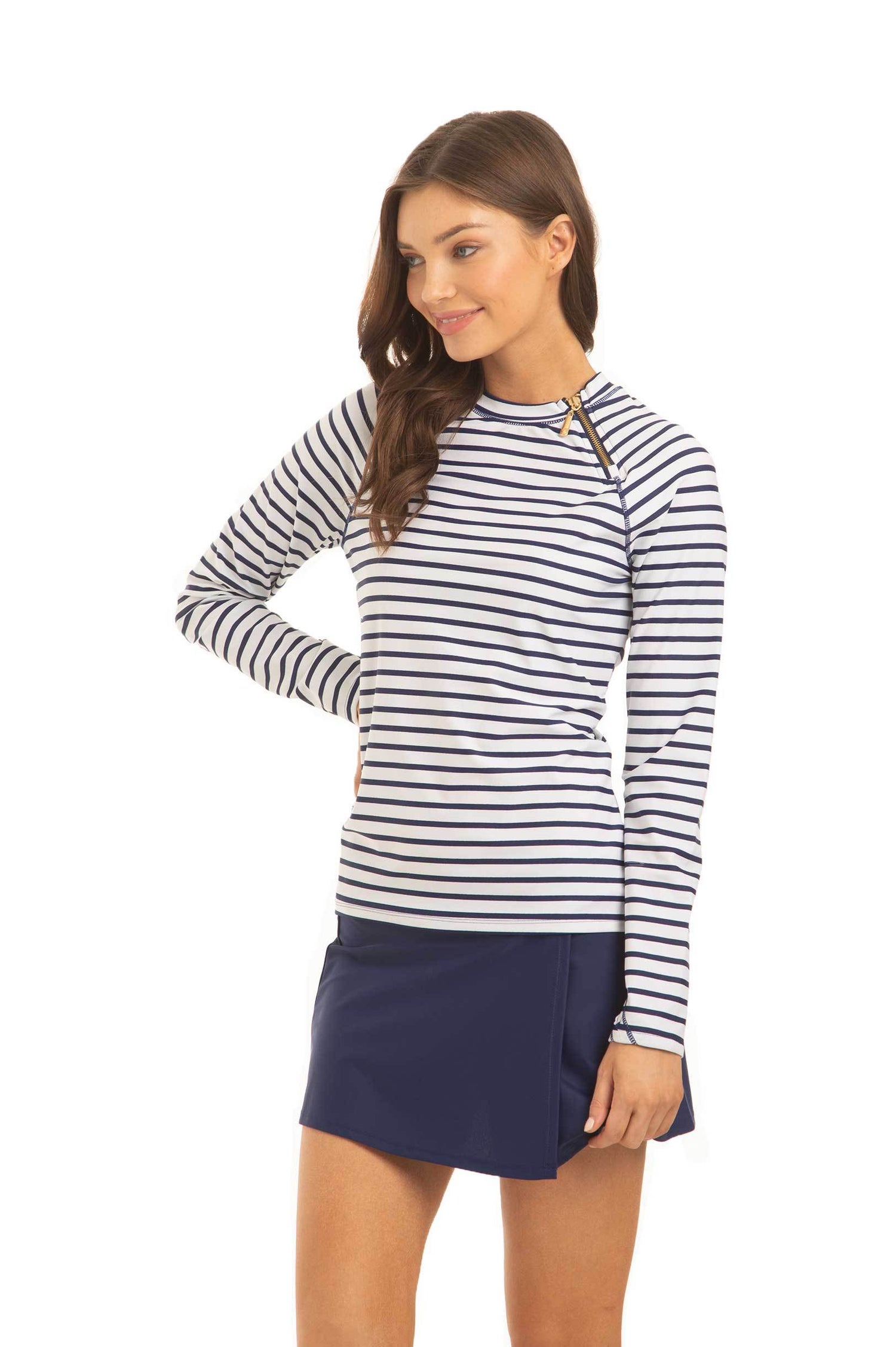 A brunette woman with her hand on her hip facing the side and wearing the Cabana Life sun protective Navy Stripe Zipper Rashguard and Navy Classic Swim Skirt.
