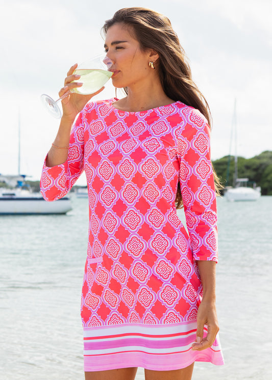 Woman in a pink patterned dress by a body of water with sailboats.
