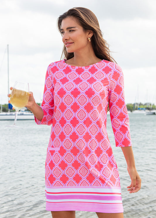 Woman in a pink patterned dress by a waterfront holding a drink.