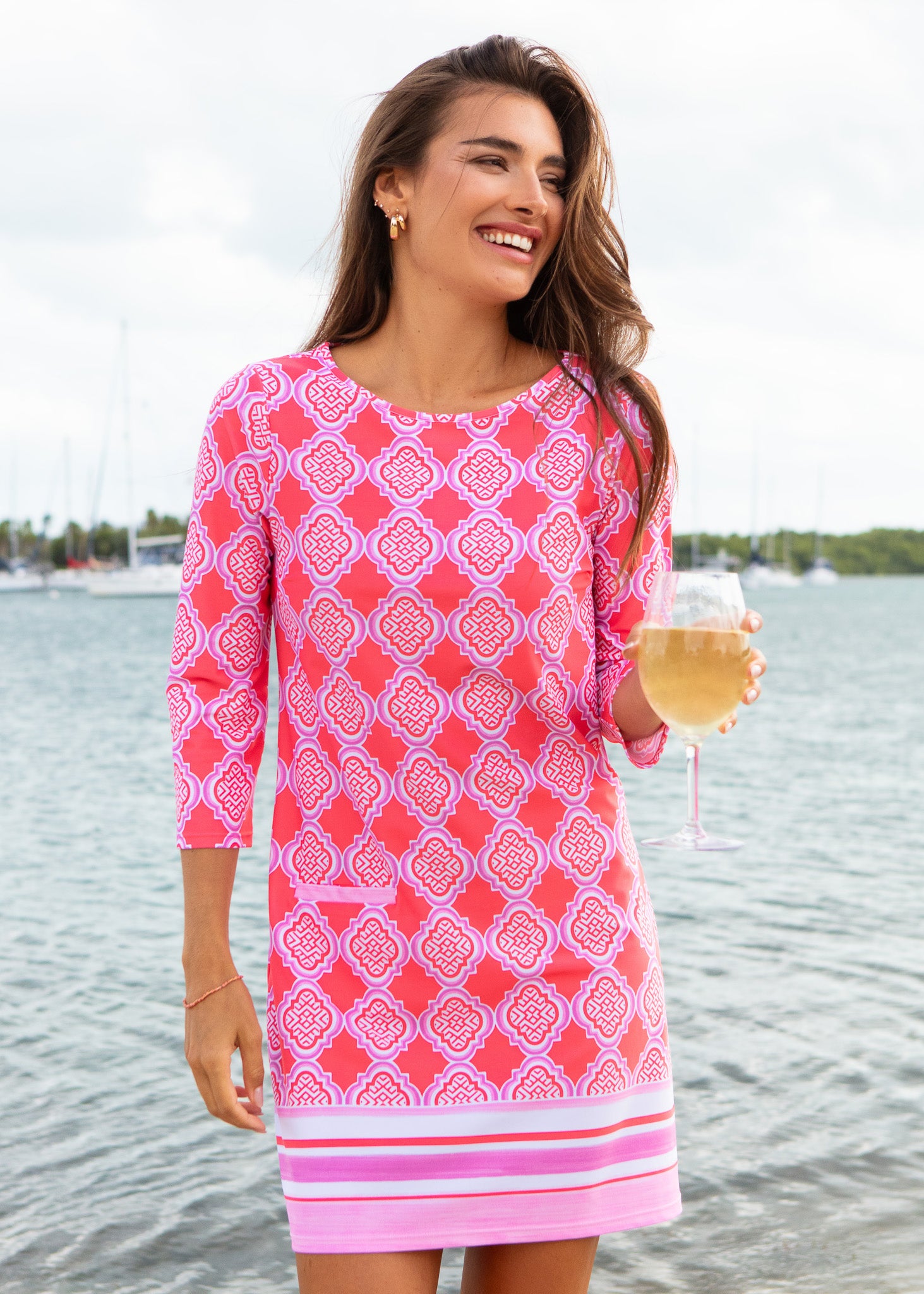 Woman in a pink patterned dress holding a glass of wine by a body of water.