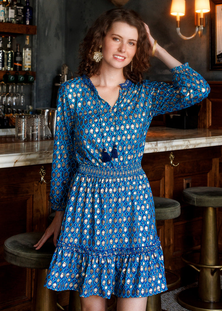 A woman wearing a blue long-sleeve dress with a gold geometric pattern, posing in front of a rustic wooden bar.