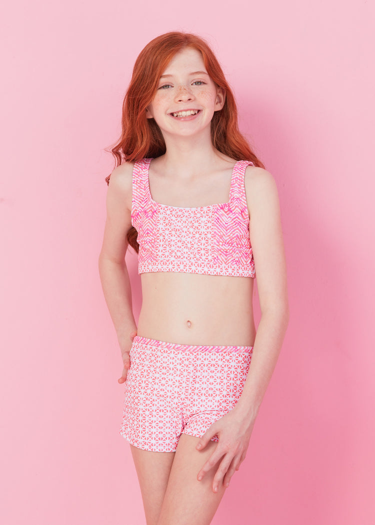 Redhead girl wearing bikini and boys short from Girls Algarve 3-Piece Long Sleeve Rashguard Set in front of pink wall