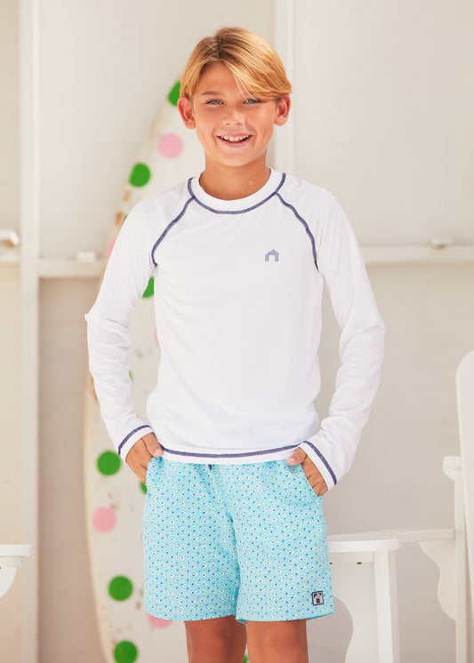 Blonde boy wearing Amalfi Coast Boys Swim Trunks, with a long sleeved white Cabana Life top.