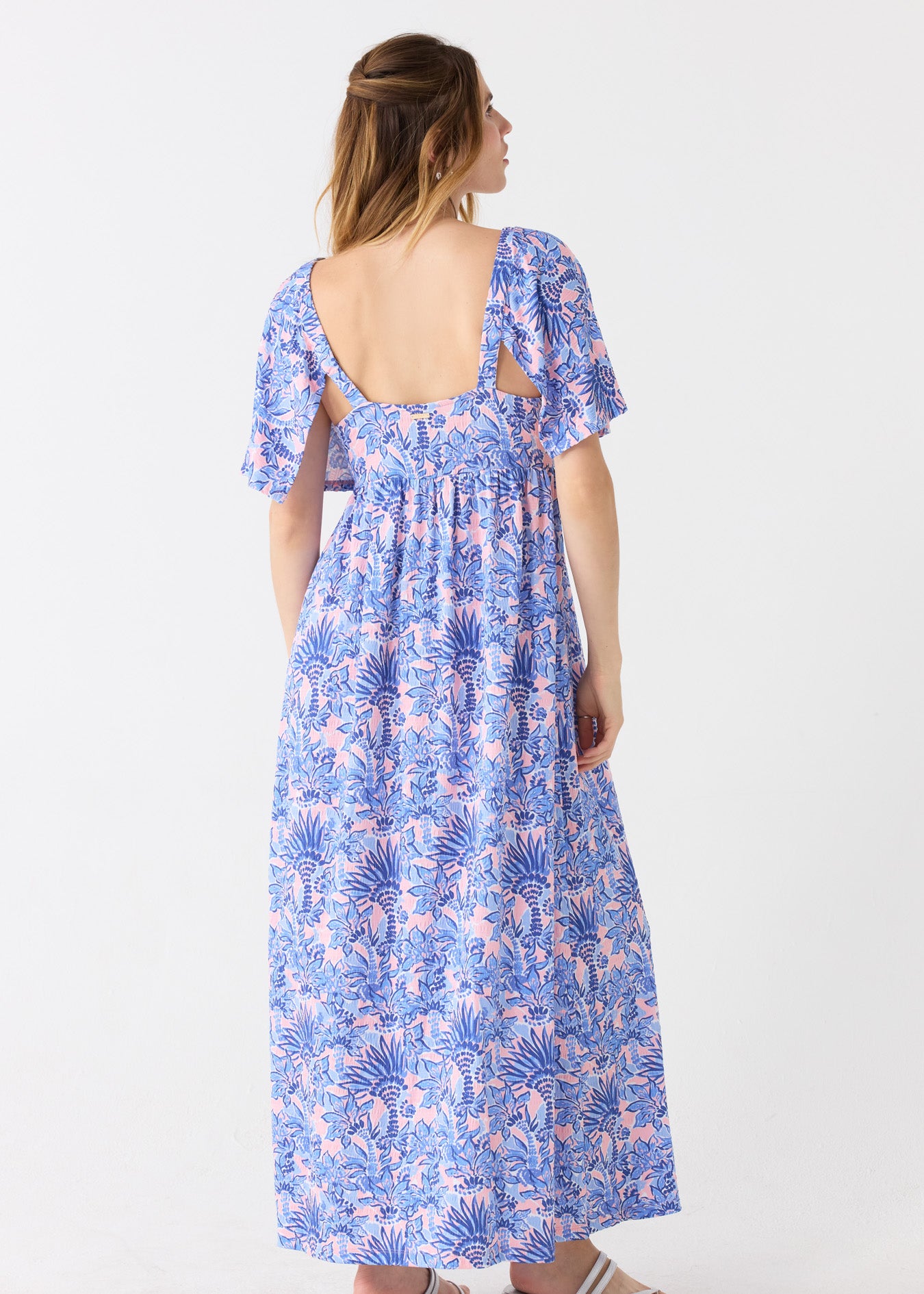 The back of a blonde woman wearing the Cabana Life sun protective Palm Beach Flutter Sleeve Maxi Dress with an open back and silver sandals.