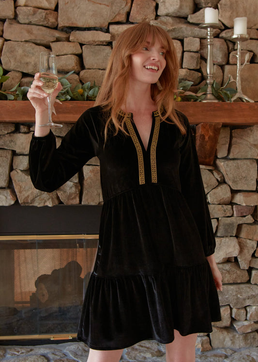 A redhead woman moving while holding a champagne flute and wearing the Cabana Life sun protective Black Embroidered Velvet Tiered Dress in front of a brick fireplace.
