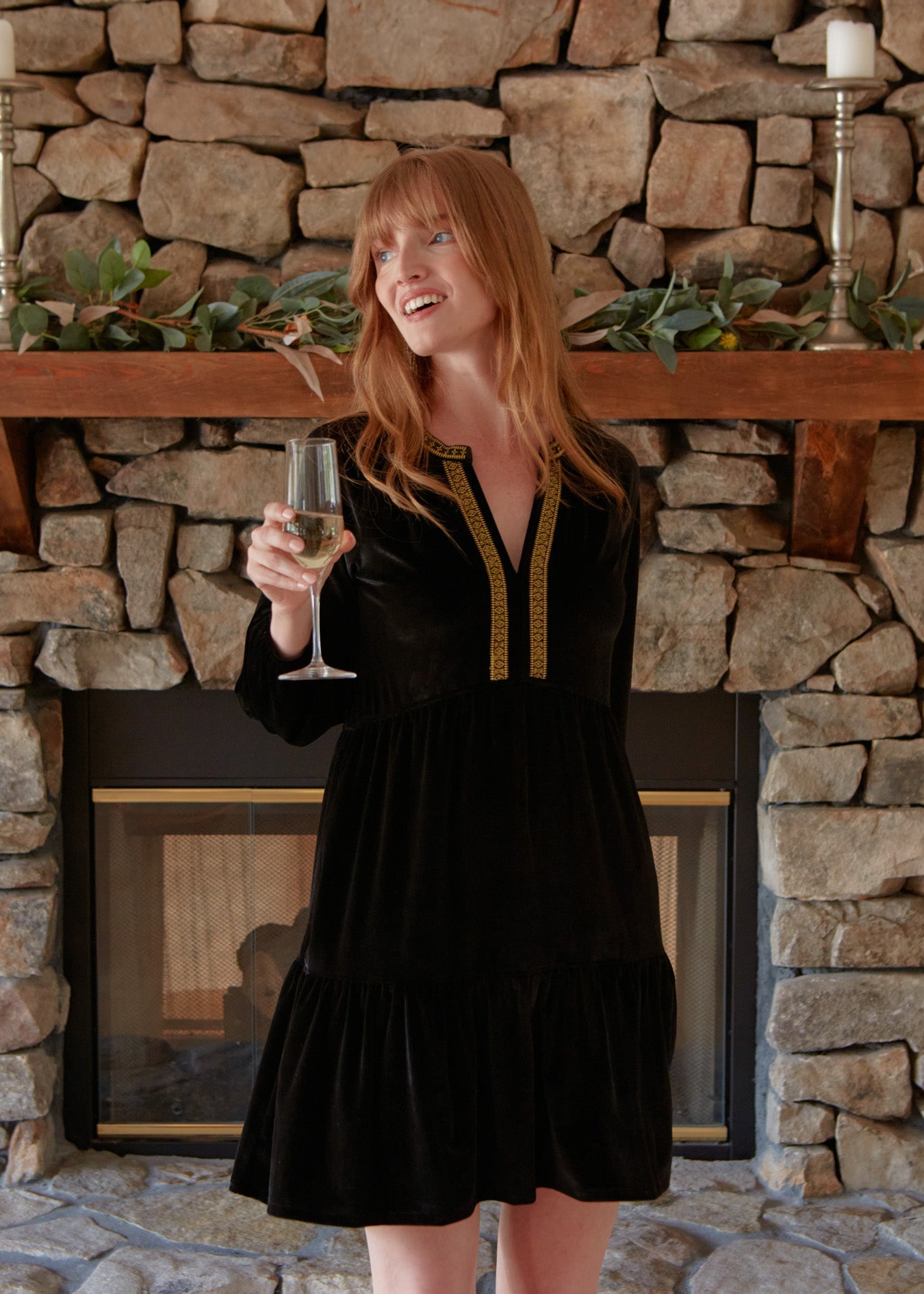 A redhead woman looking to the side and holding a champagne flute while wearing the Cabana Life sun protective Black Embroidered Velvet Tiered Dress in front of a brick fireplace.