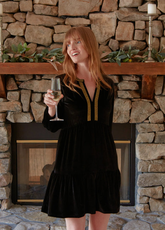 A redhead woman looking to the side and holding a champagne flute while wearing the Cabana Life sun protective Black Embroidered Velvet Tiered Dress in front of a brick fireplace.