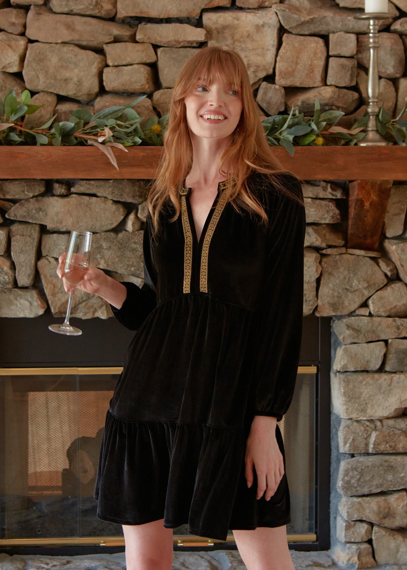 A redhead woman holding a champagne flute while wearing the Cabana Life sun protective Black Embroidered Velvet Tiered Dress in front of a brick fireplace.