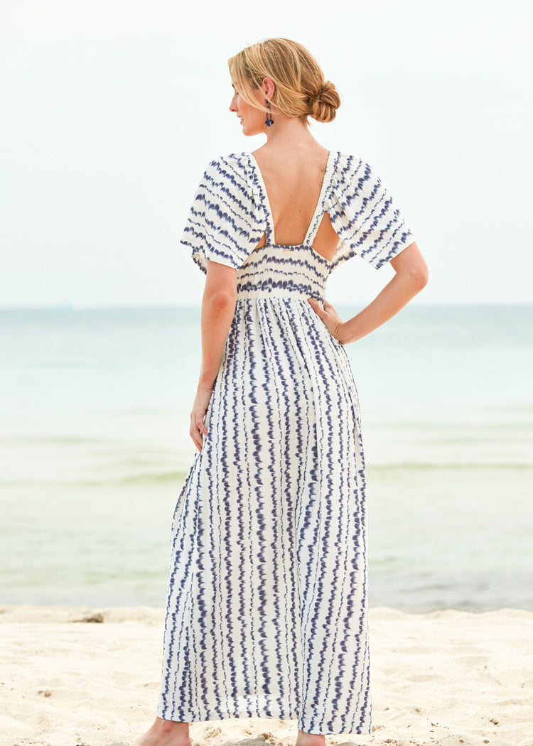 A blonde woman facing behind standing on the beach wearing the San Sebastian Flutter Sleeve Maxi Dress and navy earrings.
