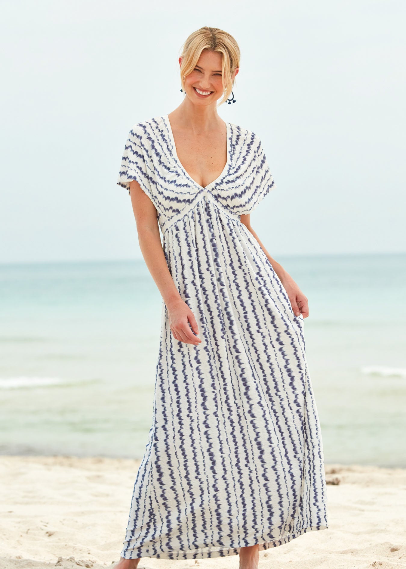 A blonde woman smiling and standing on the beach wearing the San Sebastian Flutter Sleeve Maxi Dress and navy earrings.