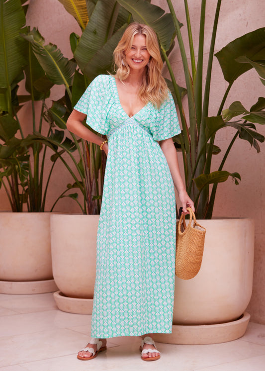 A woman with blonde hair standing in front of potted plants wearing the Cote D`Azur Flutter Sleeve Maxi Dress and holding a straw bag.