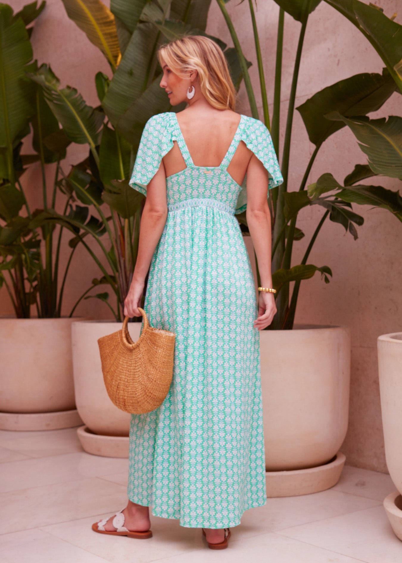 Back of a woman with blonde hair standing in front of potted plants wearing the Cote D`Azur Flutter Sleeve Maxi Dress and holding a straw bag.