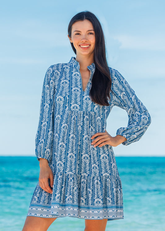 Woman wearing a blue and white patterned dress standing on a beach with clear blue water and sky.