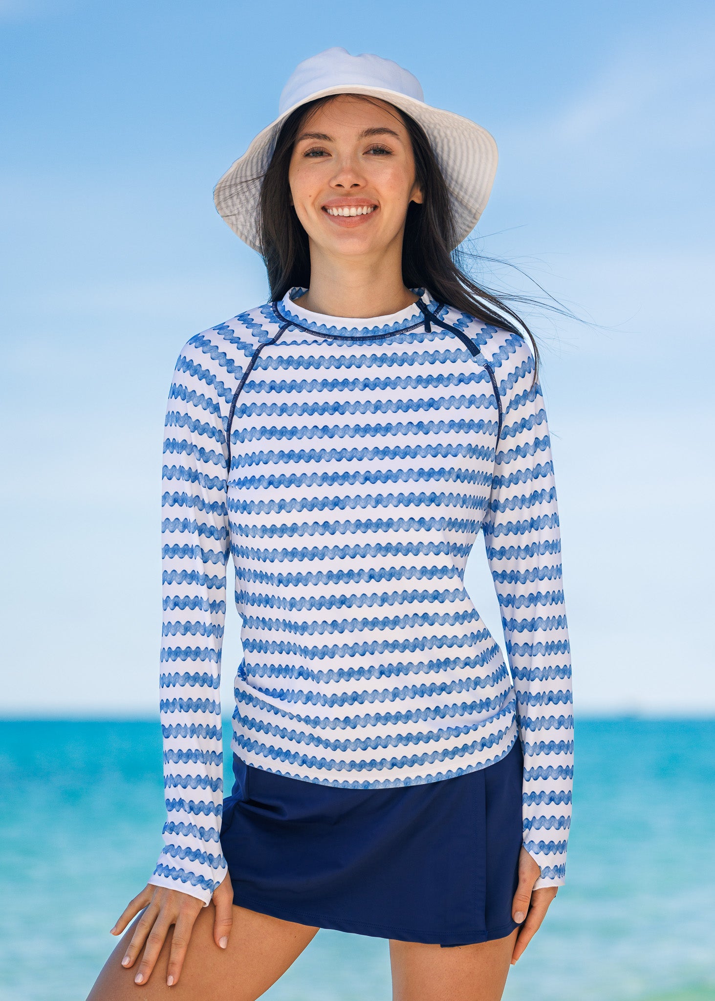 Woman wearing a blue and white rashguard with a navy swim skirt and a hat  by the ocean.