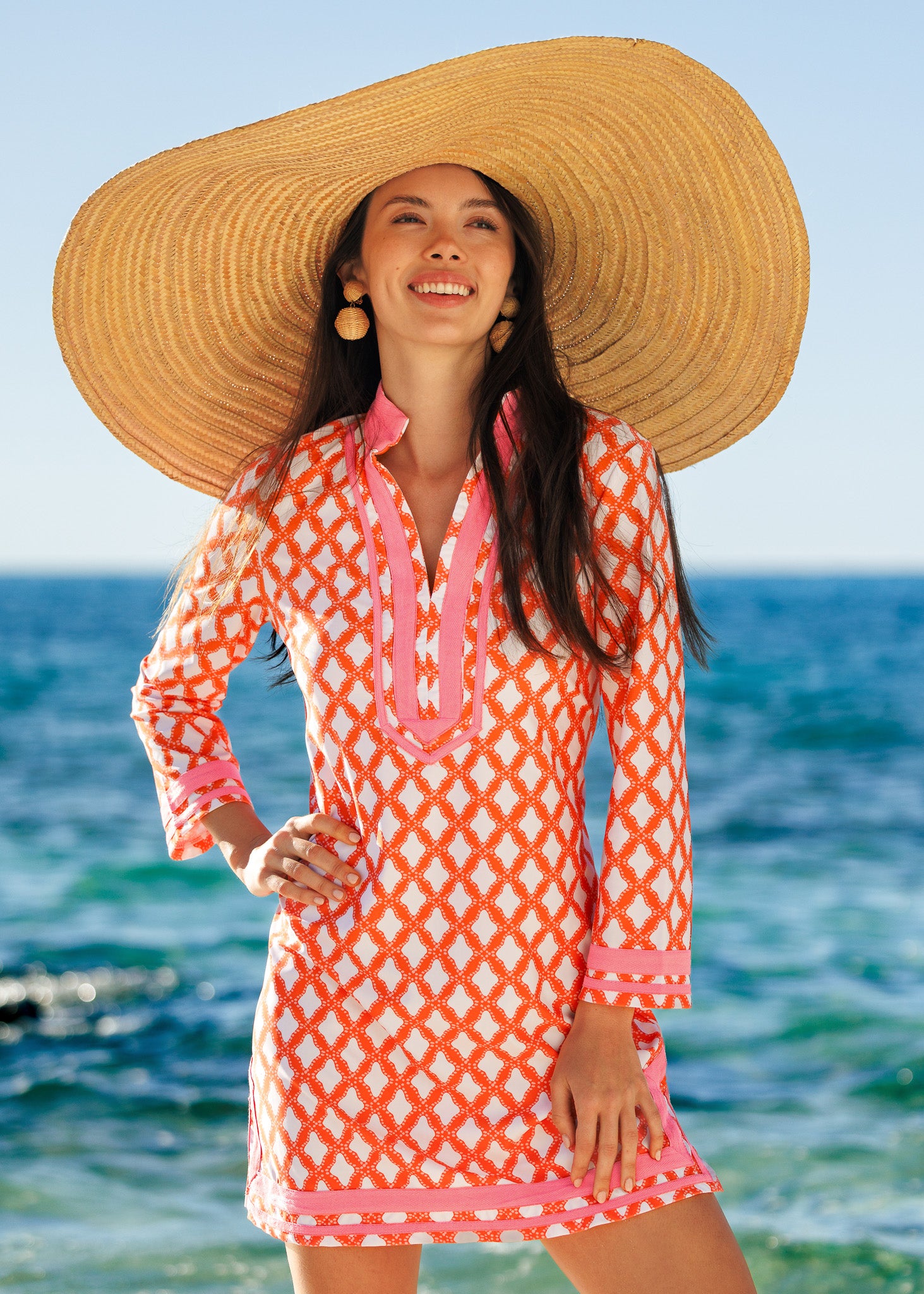 Woman in a pink and orange patterned dress with a wide-brimmed hat standing by the ocean.