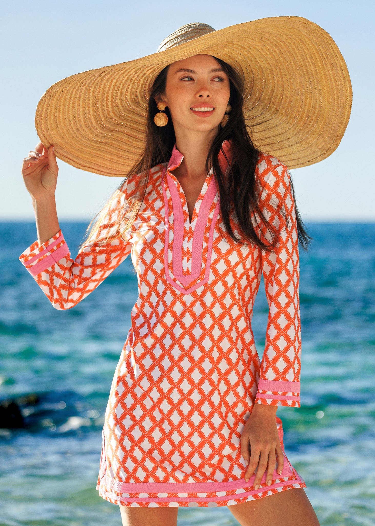 Woman wearing a large straw hat and orange patterned dress by the ocean.