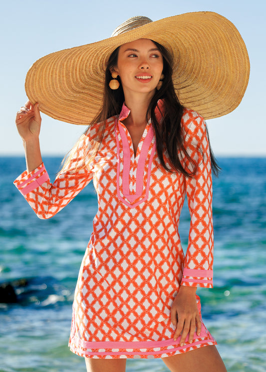 Woman wearing a large straw hat and orange patterned dress by the ocean.