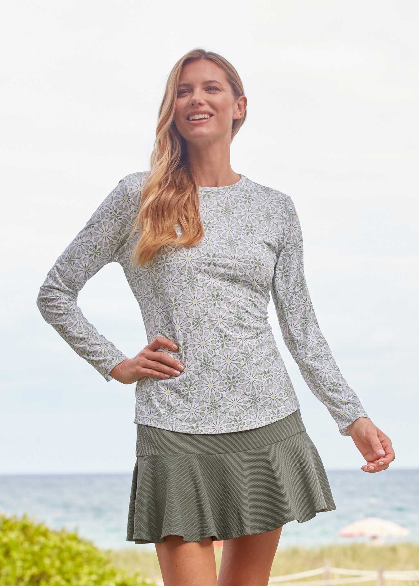 A blonde woman smiling with a hand on her hip wearing the Cabana Life sun protective Tennis Long Sleeve Performance Top and Olive Green 15" Flounce Skort.