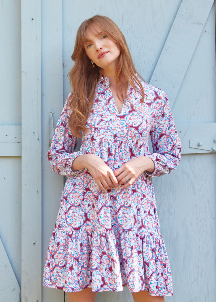 A redhead woman with her arms in front of waist while wearing the sun protective Cabana Life Camden Peony Relaxed Dress in front of a light blue door.