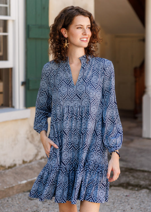 A woman wearing a blue and navy patterned long-sleeve dress with pockets, standing outdoors in front of a building with a green door.