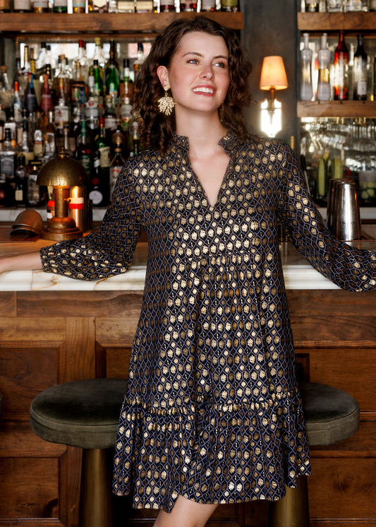 A woman wearing a long-sleeve V-neck dress with a black and gold geometric pattern, standing in a bar setting with wooden shelves and bottles in the background.