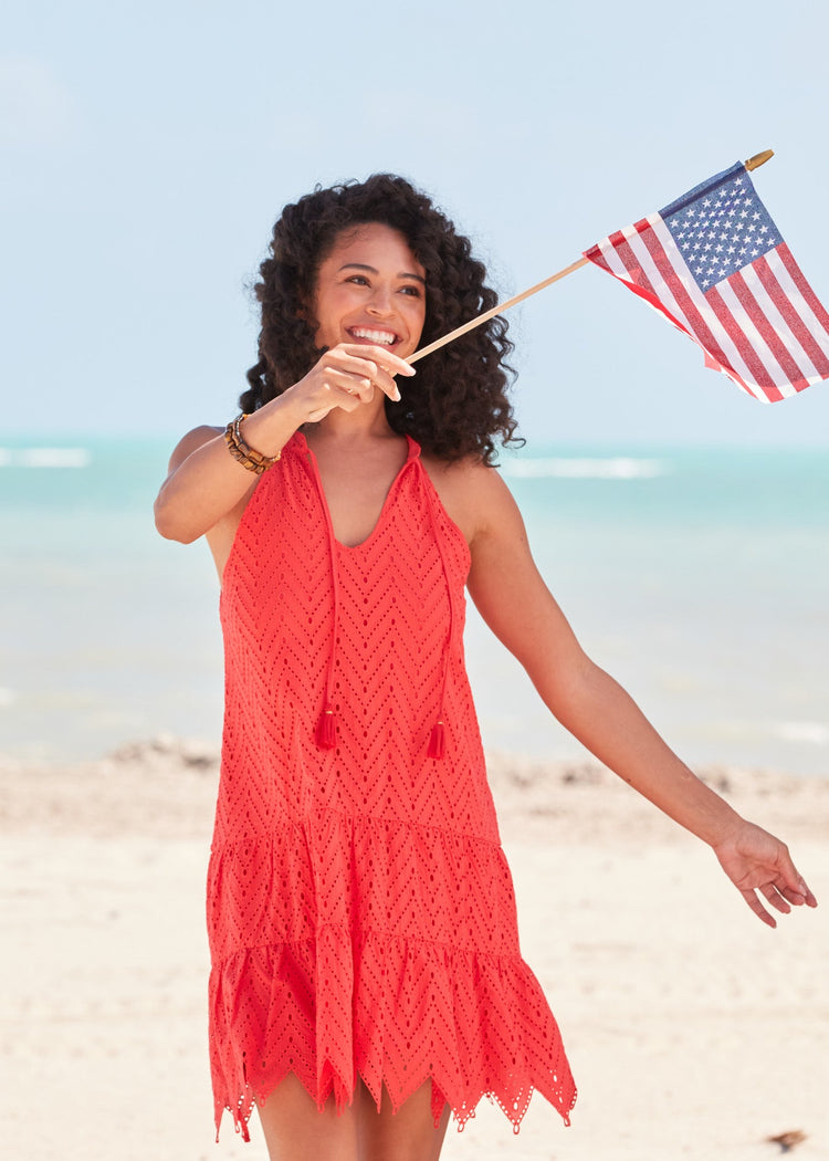 A woman with dark curly hair holding an American flag while wearing the Poppy Red Eyelet Halter Dress on the beach.