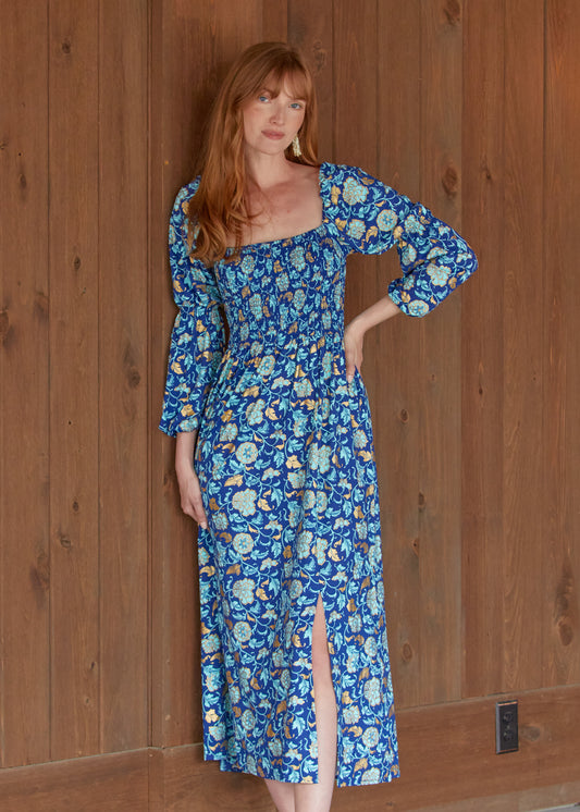 A redhead woman wearing the sun protective Navy Floral Metallic Puff Sleeve Maxi Dress from Cabana Life in front of a wood background.