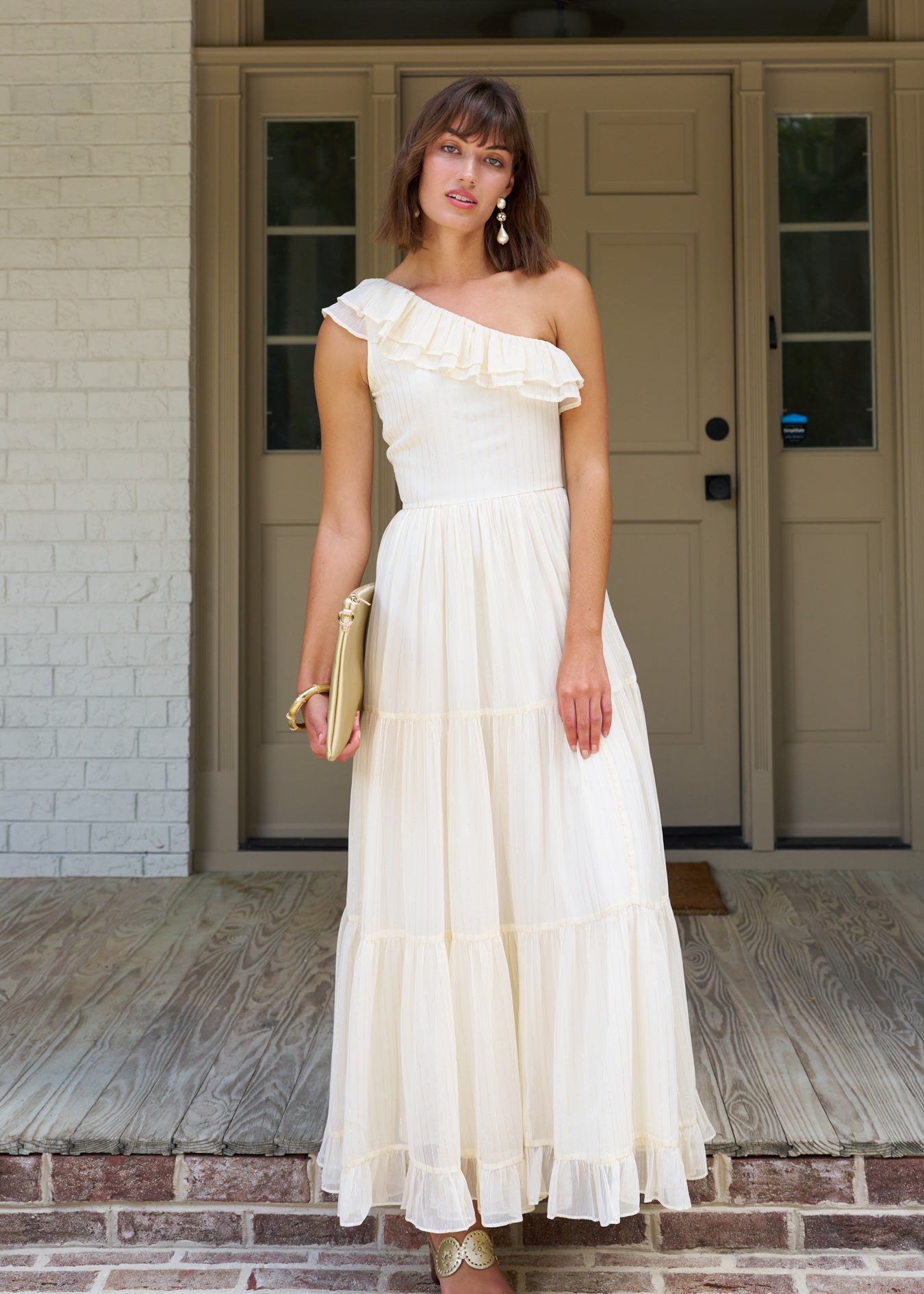 Woman with short brown hair standing on a porch wearing the ivory and gold metallic one shoulder maxi dress.