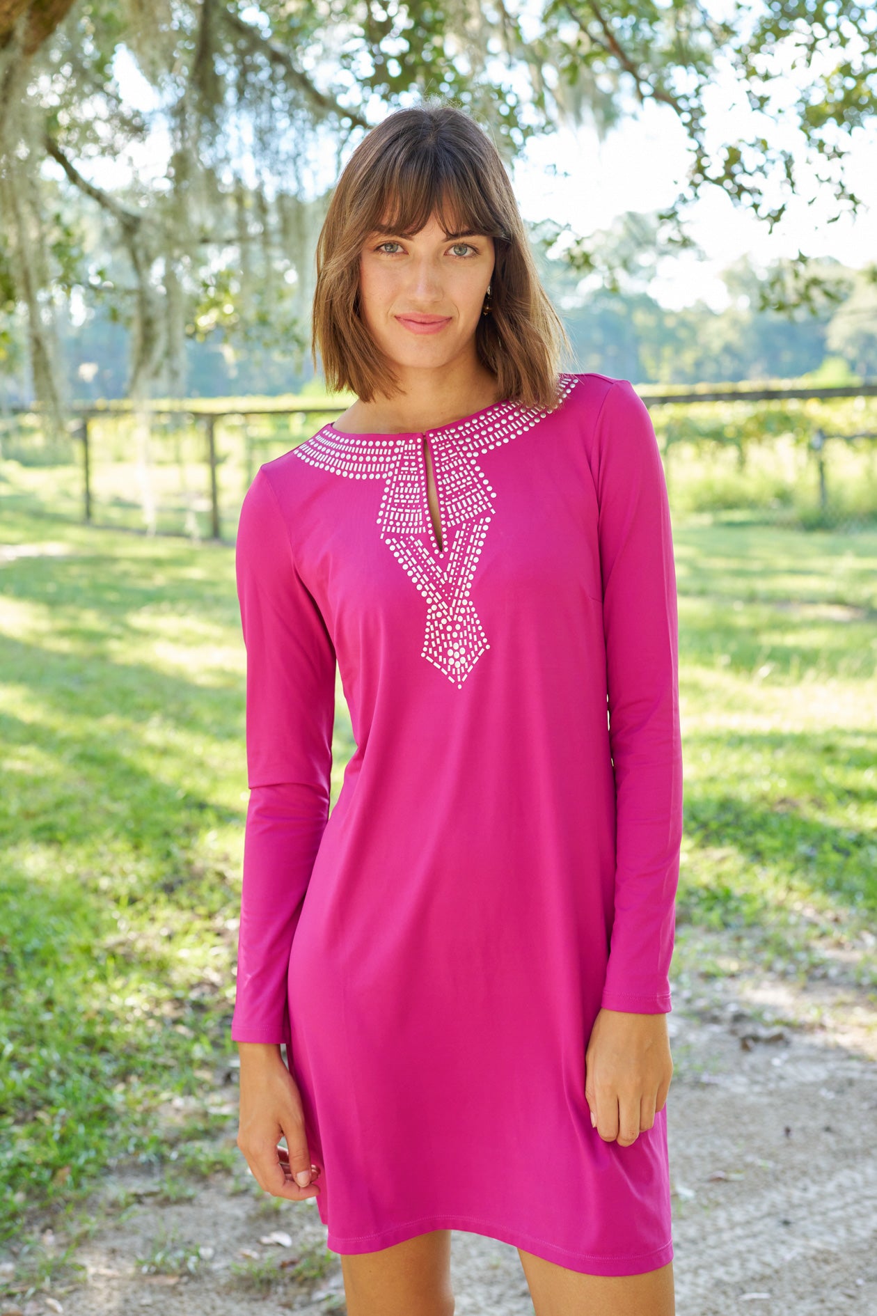 Brunette woman with short hair and bangs wearing Merlot Metallic Key Hole Dress.on a dirt road in front of a grassy field.