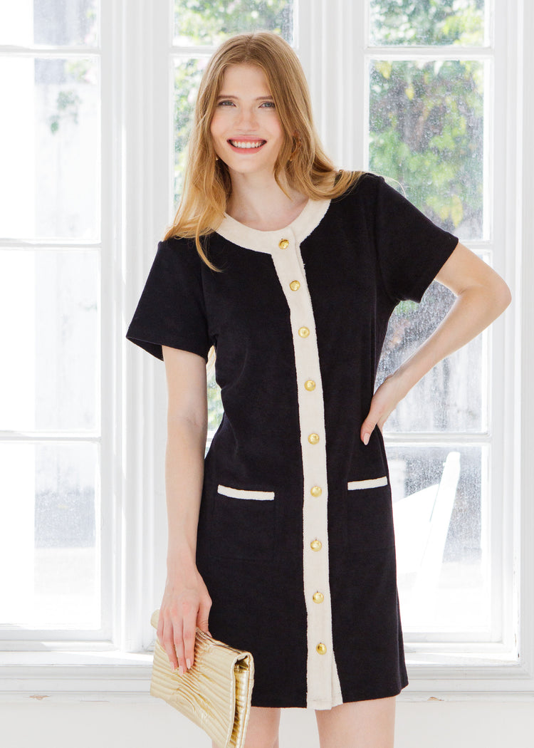 Woman wearing a black and cream terry dress with gold buttons standing outside a white window. 
