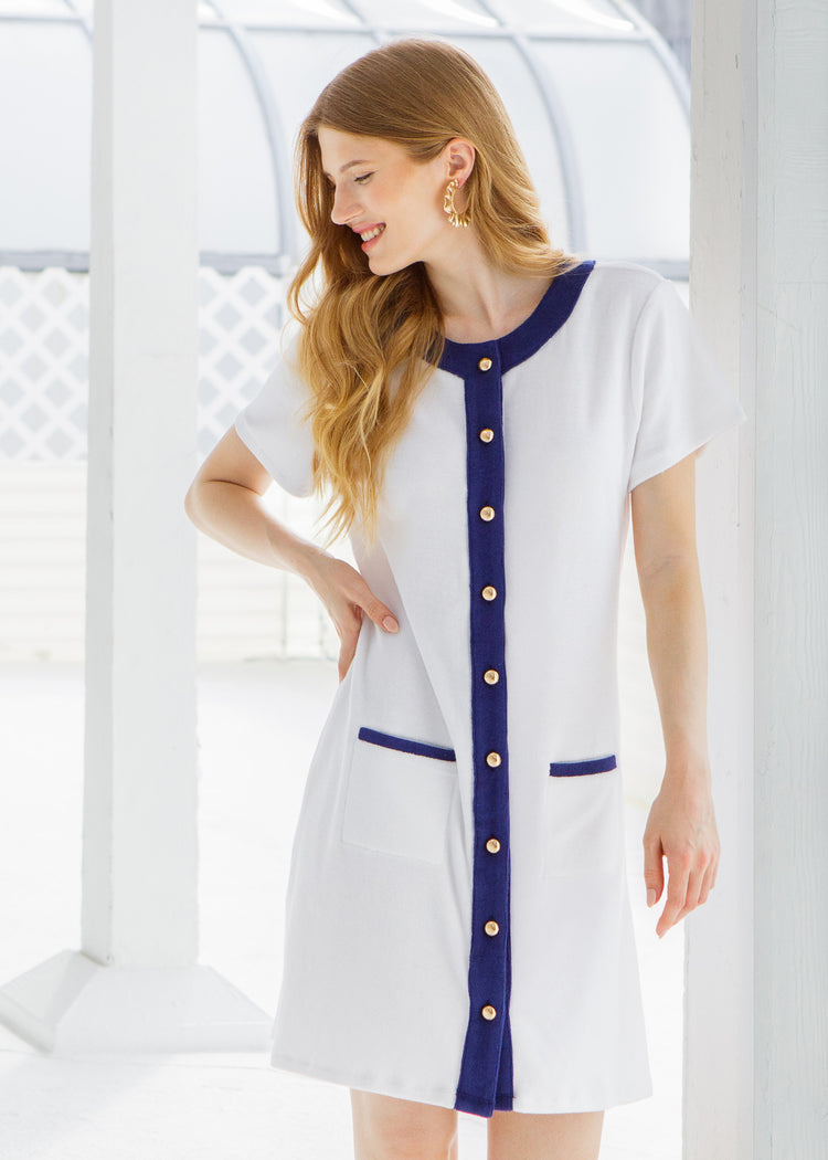 Woman wearing a white and blue terry dress with gold buttons standing outside a white cottage. 