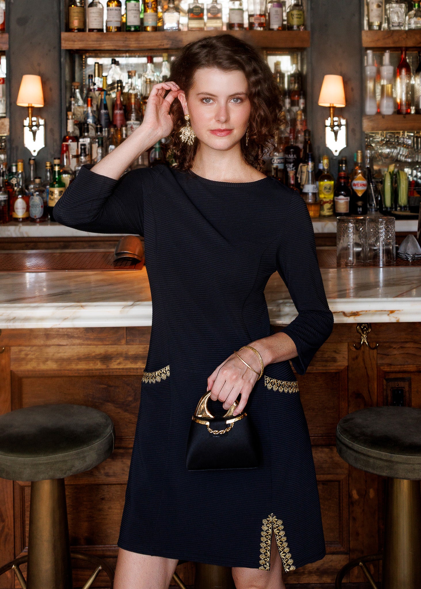 A woman stands in a bar setting wearing a black knee-length dress with gold embroidered detailing at the waist and hem, holding a matching small black handbag with a gold ring handle.