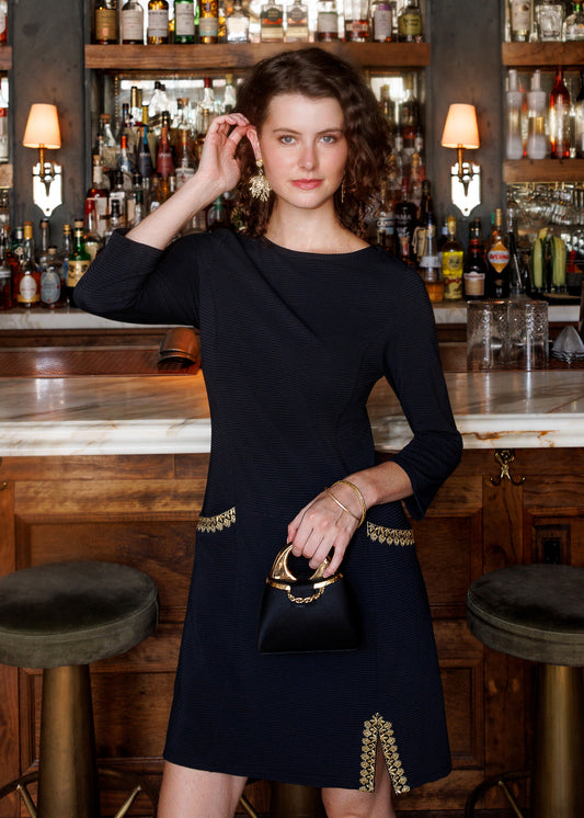 A woman stands in a bar setting wearing a black knee-length dress with gold embroidered detailing at the waist and hem, holding a matching small black handbag with a gold ring handle.
