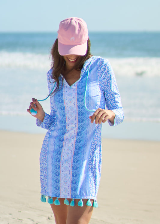 Woman looking down wearing Lake Como Hooded Tunic Dress with Pink Cabana Life Baseball Hat holding tassels on beach.