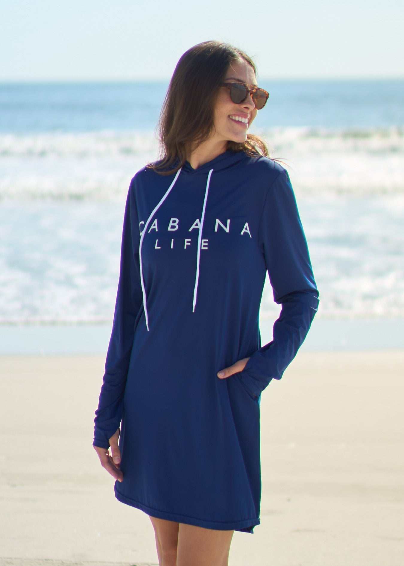 A woman smiling and looking to the side wearing the Navy Cabana Life Hoodie Dress with hand in pocket on the beach.