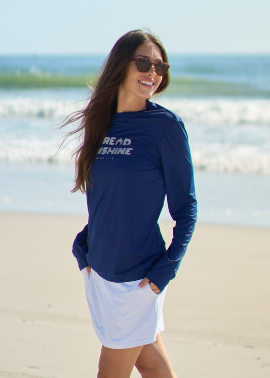 A woman smiling and looking to the side wearing the Navy Spread Sunshine Unisex Performance Shirt with hands in pockets of the White Skort on the beach.