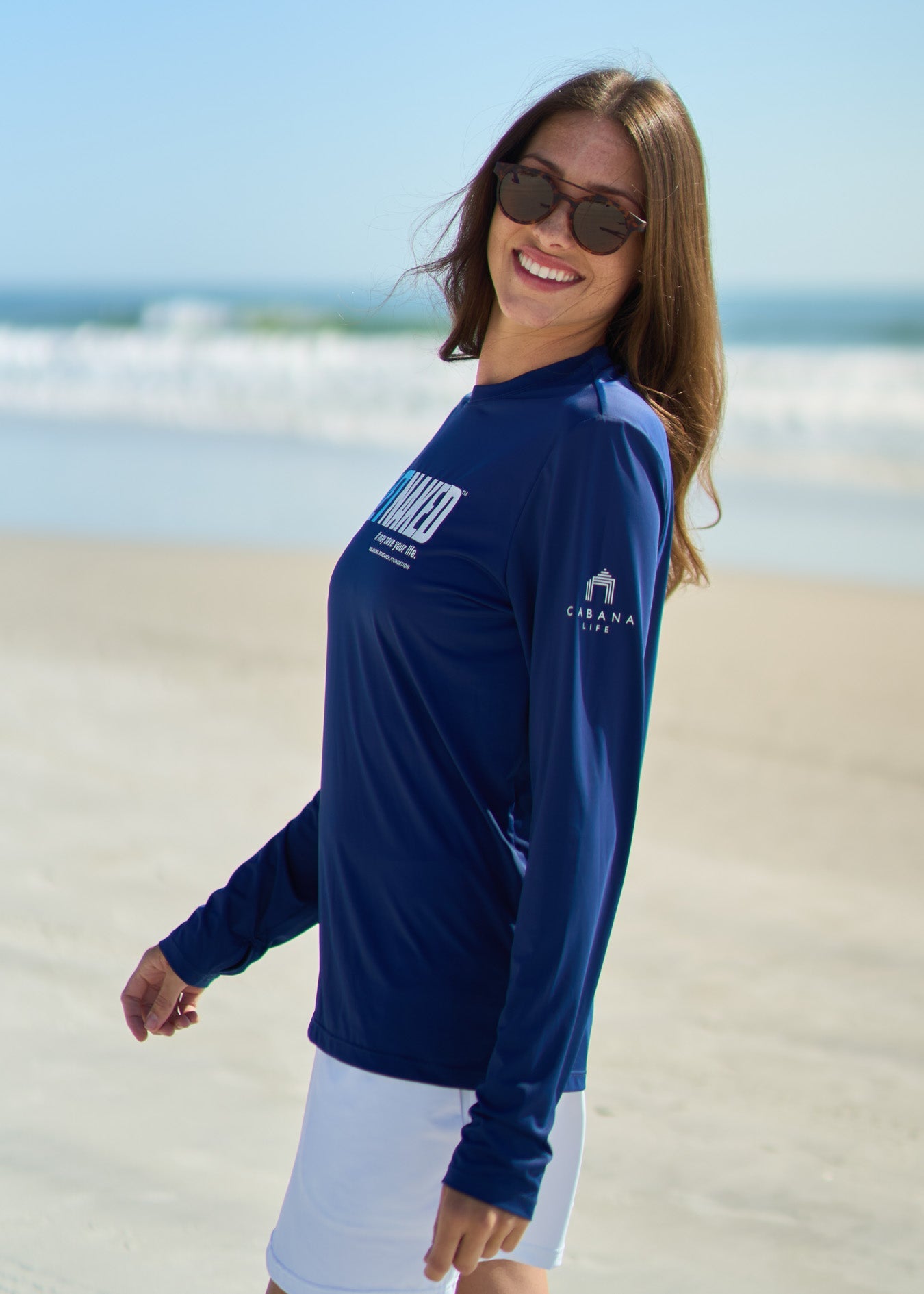 A woman facing to the side on the beach wearing the Get Naked Unisex Performance Shirt and White Skort.