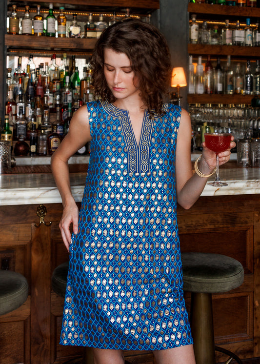 A woman stands in a bar setting wearing a sleeveless blue dress with a gold geometric pattern and embroidered neckline. She is holding a red cocktail glass, with wooden bar stools and shelves of liquor bottles in the background.