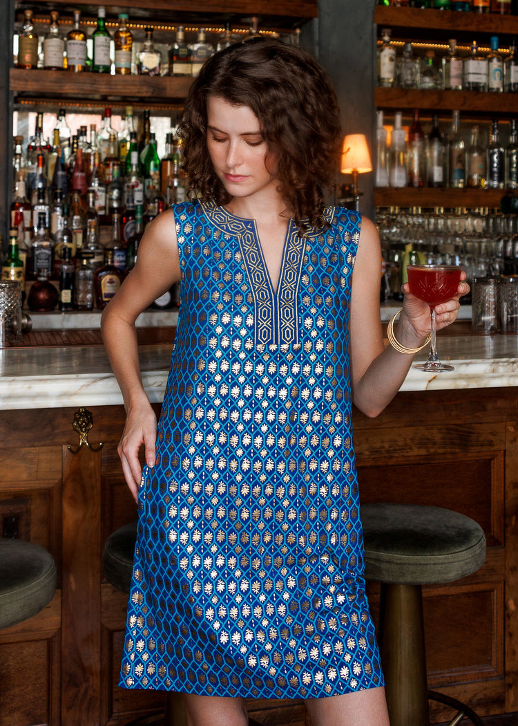 A woman stands in a bar setting wearing a sleeveless blue dress with a gold geometric pattern and embroidered neckline. She is holding a red cocktail glass, with wooden bar stools and shelves of liquor bottles in the background.