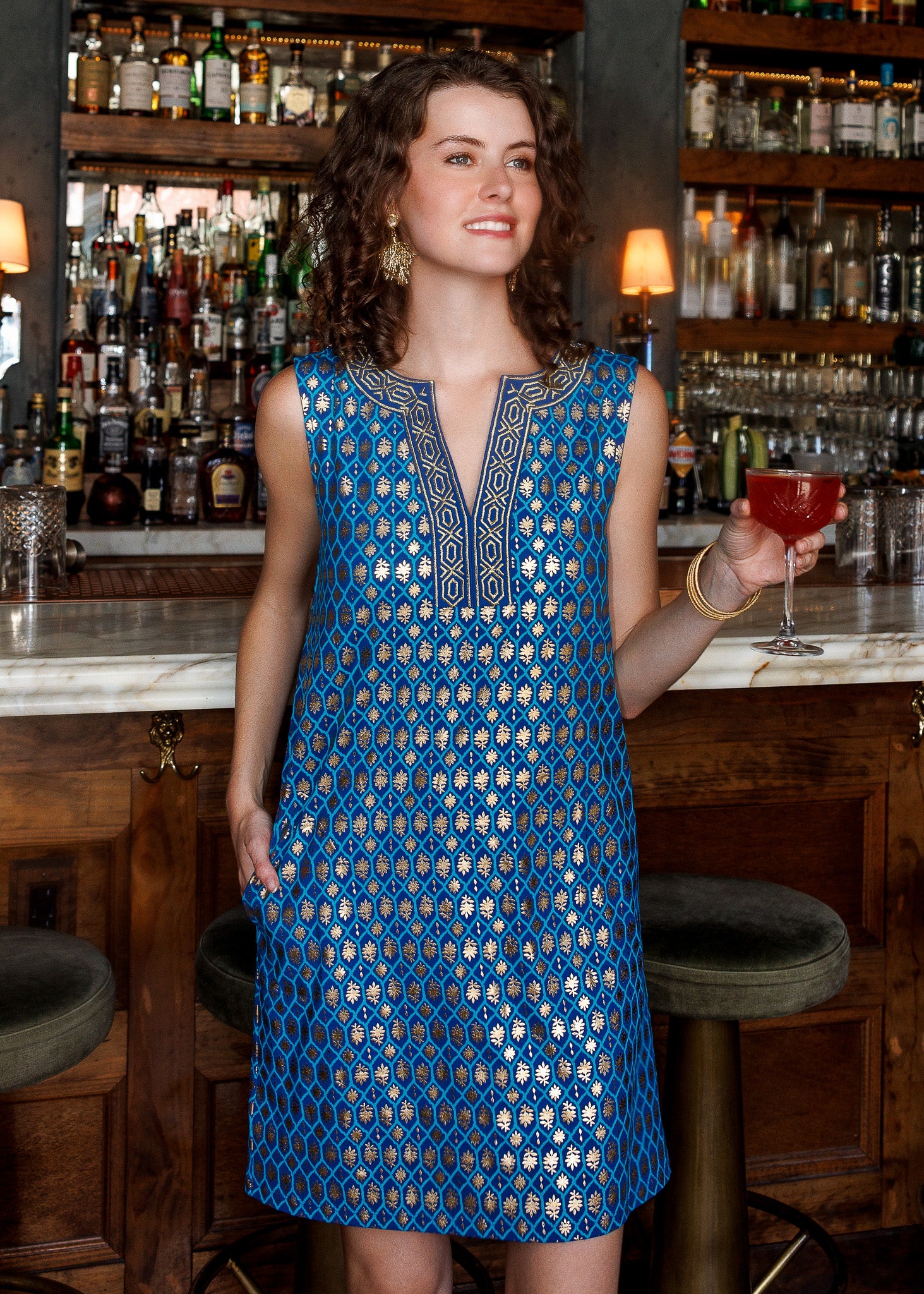 A woman stands in a bar setting wearing a sleeveless blue dress with a gold geometric pattern and embroidered neckline. She is holding a red cocktail glass, with wooden bar stools and shelves of liquor bottles in the background.