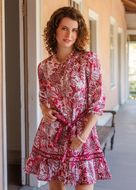 A woman with curly brown hair stands on a porch wearing a pink and white floral print mini dress with a tie waist and ruffled hem.