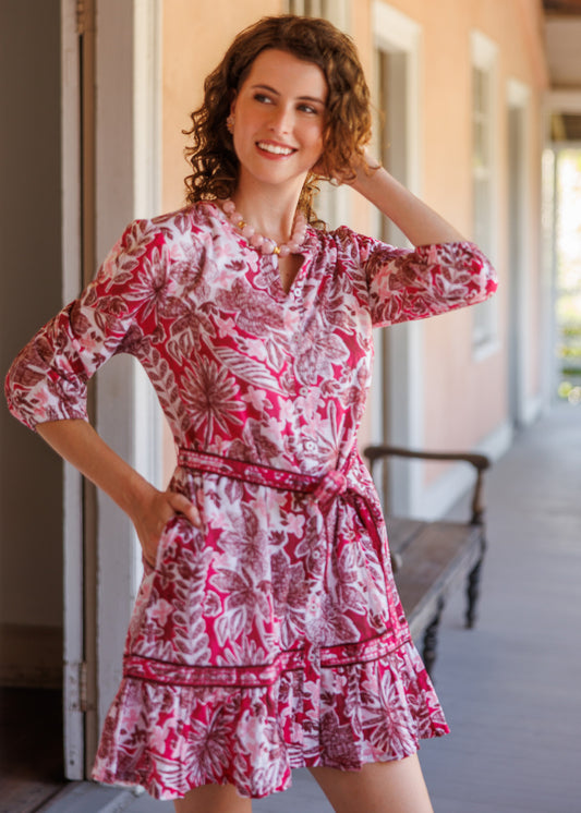 A woman modeling a pink and white floral print mini dress with three-quarter sleeves and a matching tie belt, standing on a porch.