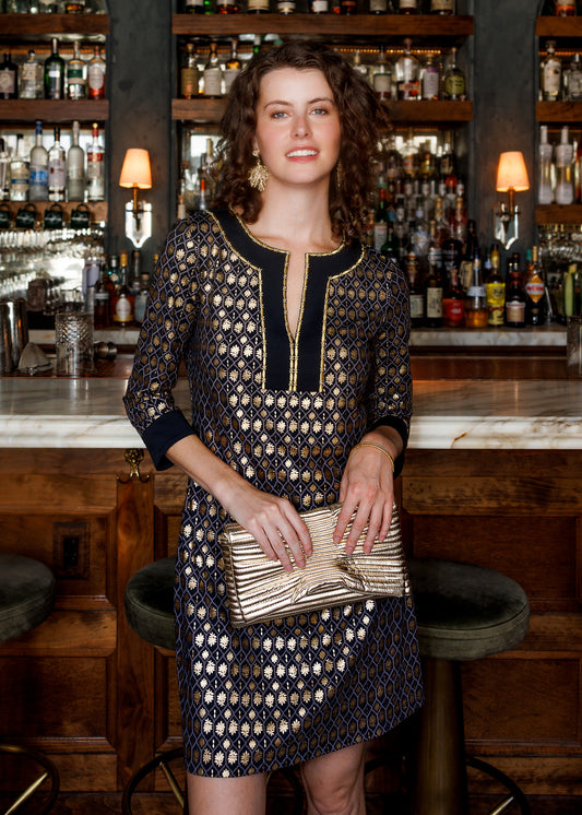 A woman stands in a bar setting wearing a navy blue and gold patterned dress with a gold clutch bag.