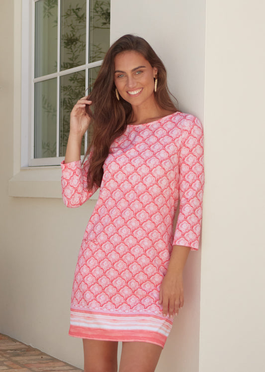 Woman wearing a pink patterned dress standing against a white wall.