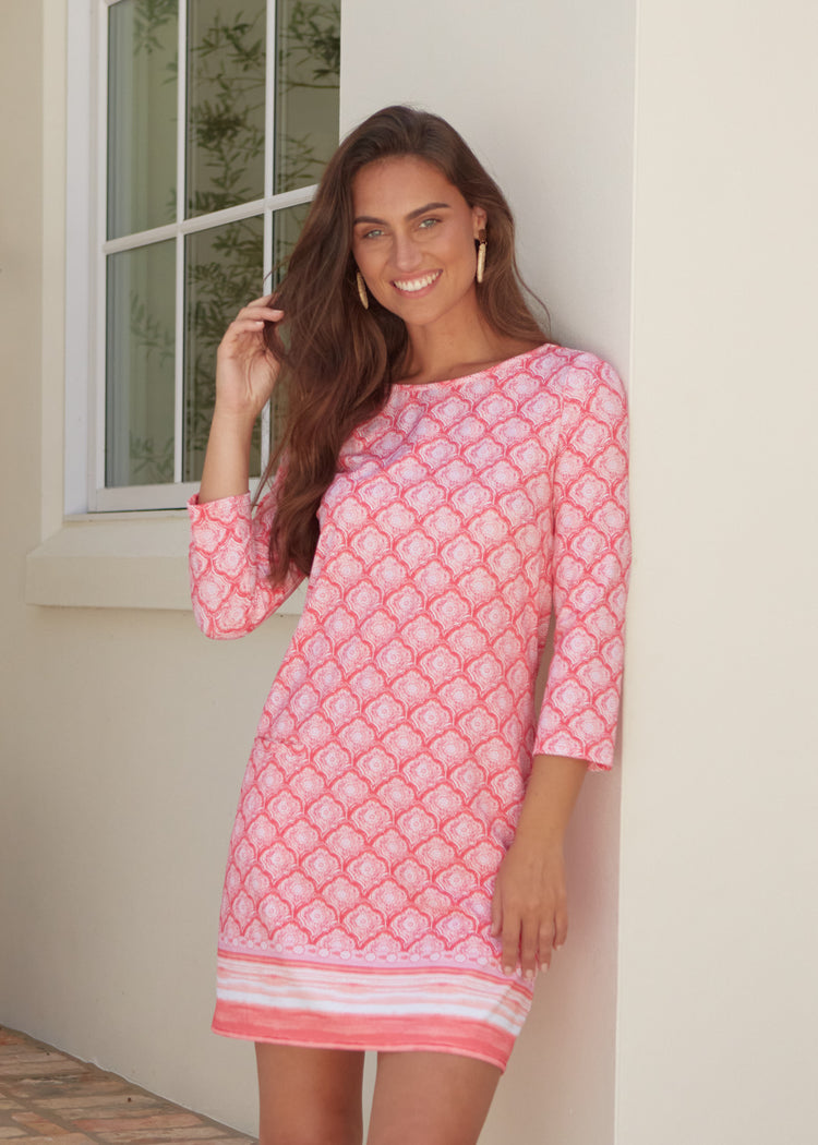 Woman wearing a pink patterned dress standing against a white wall.