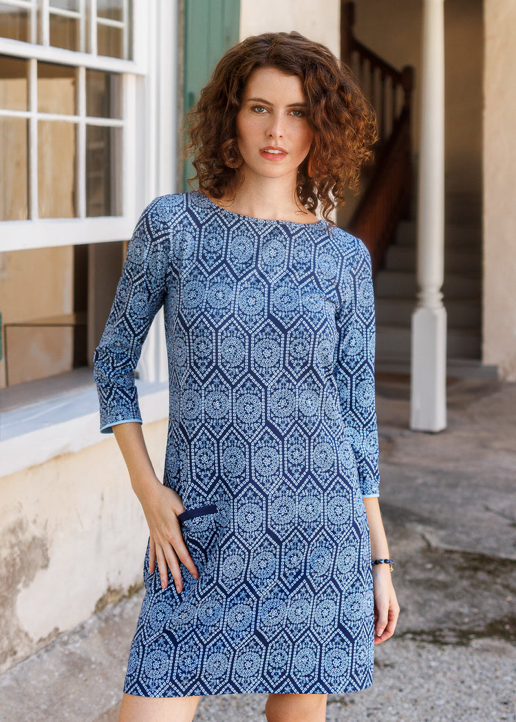 A woman wearing a blue and navy patterned three-quarter sleeve dress, standing in front of a building with a staircase and window.